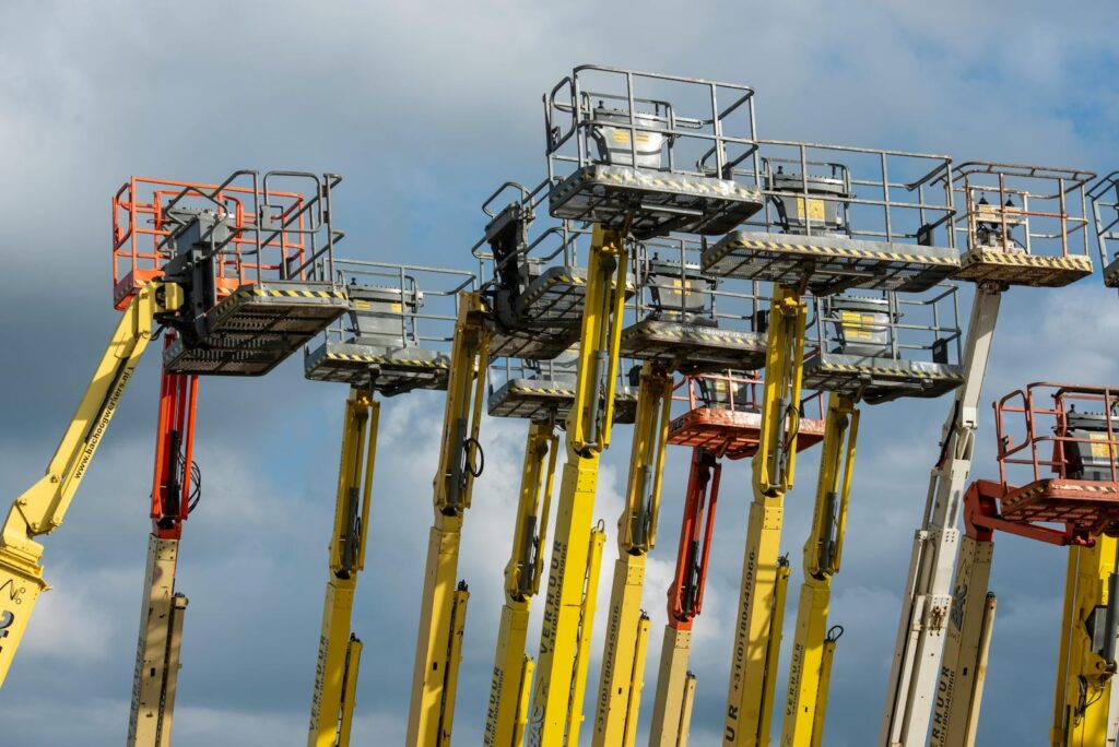 Multiple aerial work platforms with colorful booms reaching skyward against a cloud-dotted backdrop.